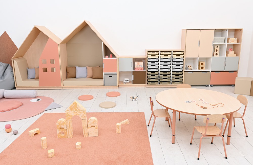 Ljus förskolemiljö med trähus, förvaringsmöbler, runda bord och mjuka pastellfärger i rosa och beige toner.
