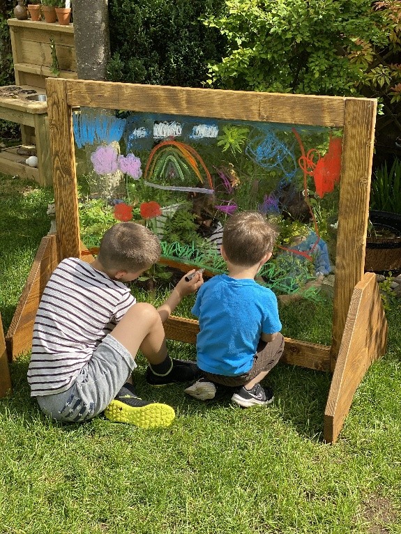 Barn målar med färg på en genomskinlig tavla i trädgården under en kreativ utomhusaktivitet.